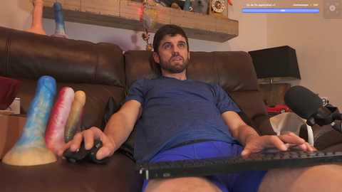 Media: Video of a man with short brown hair and beard, wearing blue shorts and a dark blue t-shirt, playing video games on a couch, surrounded by colorful sex toys and a microphone.