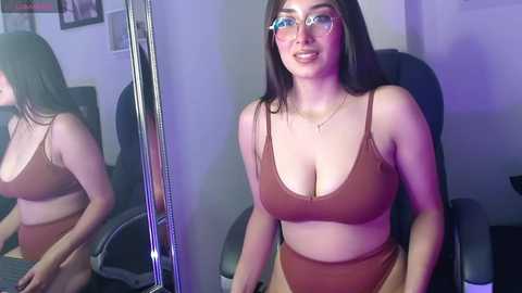 Media: Video of a curvy Latina woman with long dark hair, wearing glasses and a brown bikini, sitting in a dark office chair, with a mirror reflecting her image.