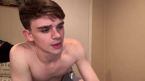 Media: Video of a young, shirtless, fair-skinned boy with short brown hair, looking serious in a beige room with a cluttered bed in the background.