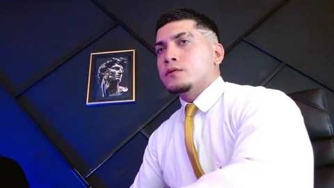 Media: Video of a young man with short dark hair and a neatly trimmed beard, wearing a white dress shirt and yellow tie, standing in a modern office with dark walls and a black-and-white framed portrait.