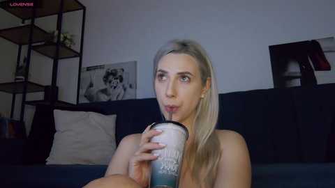 Media: Video of a blonde woman with fair skin, medium build, topless, drinking from a glass bottle on a dark blue couch. Background features black shelves and framed artwork.