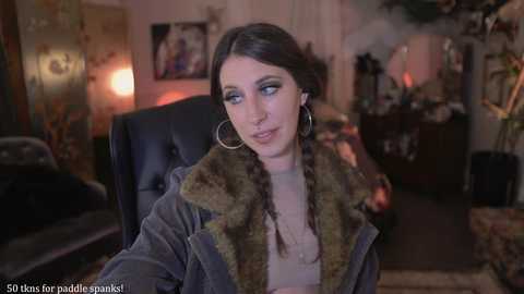 Media: Video of a young woman with dark hair in braids, wearing a fur-lined jacket, and hoop earrings, sitting in a dimly lit, cozy room with eclectic decor.