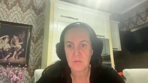 Media: Video of a middle-aged woman with dark hair, wearing a black jacket, speaking in a living room with patterned wallpaper, framed artwork, and modern kitchen cabinets in the background.