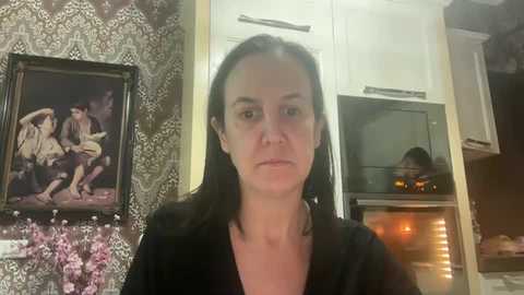 Media: A video of a woman with straight black hair, wearing a black top, standing in a kitchen with floral wallpaper, framed art, and a lit oven.