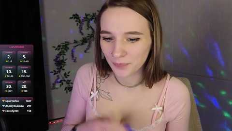 Media: Video of a fair-skinned woman with straight brown hair, wearing a pink camisole, sitting in a dimly lit room with a purple and blue light display.