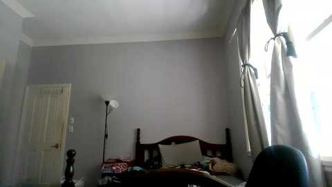 Media: Video of a sparsely furnished, dimly lit bedroom with a wooden bed, beige curtains, and a tall floor lamp.