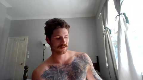 Media: Video of a shirtless, bearded man with a large, colorful tattoo on his chest, standing in a brightly lit bedroom with grey walls and white curtains.