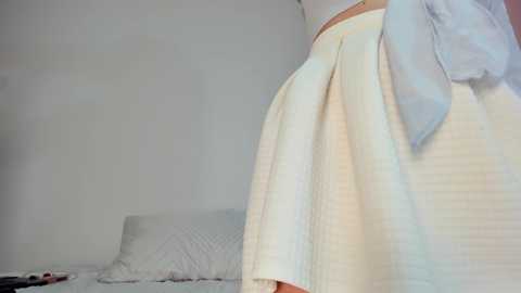 Media: Video of a woman in a white dress with a pleated skirt, standing in a minimalist, white bedroom with a bed, pillows, and a small table.