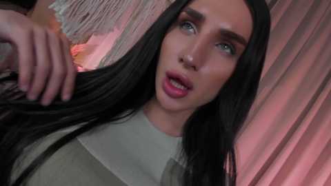 Video of a young woman with long black hair, light skin, and blue eyes, wearing a light-colored top, against a pink, draped background. She looks surprised and slightly open-mouthed.