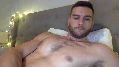 Media: Video of a muscular, bearded, shirtless man with a light complexion lying on a bed with white pillows and a gray headboard. Background features a string of warm, yellow fairy lights and a subtle, textured wall.