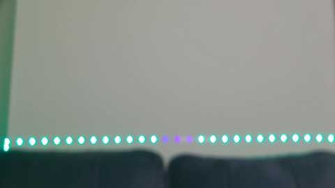 Media: A video of a dimly lit room showing a wall-mounted string of green LED lights with a faint purple hue, positioned horizontally across the top of the image. The background is a plain, light gray wall, and the bottom half features a dark blue, plush sofa.