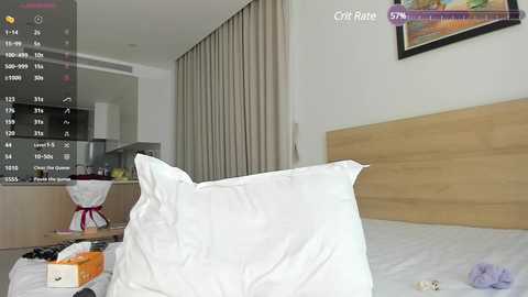 Media: Video of a modern bedroom with a large wooden headboard, white pillows, and a messy bed with clothes and a tissue box. The background features beige curtains and a framed painting.
