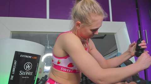 Media: Video of a blonde woman in a pink sports bra and shorts, working out with a black dumbbell in a gym with purple walls and white trim.