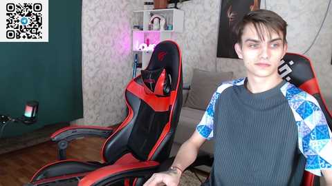 Media: Video of a young man in a gaming chair, wearing a blue and black shirt, in a dimly lit room with a green wall and a QR code in the corner.