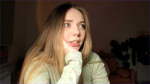 Media: Video of a young woman with long, light brown hair, wearing a light green pajama top, gazing thoughtfully with her hand on her chin, in a dimly lit room with a potted plant and a bed in the background.