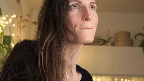 Media: Video of a young woman with long, straight, dark brown hair and fair skin, looking contemplative. She wears a black top. Background includes a potted plant, warm yellow string lights, and beige walls.