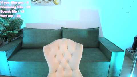 Media: Video of a green, tufted couch in a brightly lit room with a white wall and potted fern. A woman in a white dress sits in the foreground. Text overlays the image.