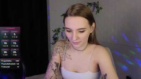 Media: Video of a young woman with light skin and brown hair, wearing a white tank top, showcasing detailed tattoo sleeves. She sits indoors with dim lighting, displaying a livestream overlay showing follower counts and social media icons.