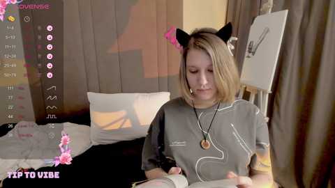 Media: A video of a young woman with light skin and blonde hair, wearing cat ears and a grey T-shirt, sitting at a desk in a cozy, dimly-lit room.