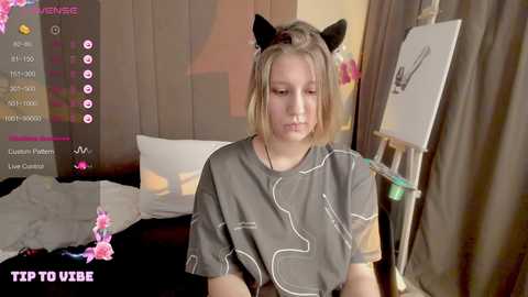 Media: A video of a young blonde girl in a gray hospital gown with cat ears, standing near a painting easel in a dimly lit room.