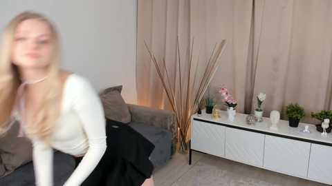 Media: Video of a blonde woman in a white cardigan and black skirt, blurred, standing in a modern living room with beige curtains, a gray sofa, a white console table, and decorative plants.