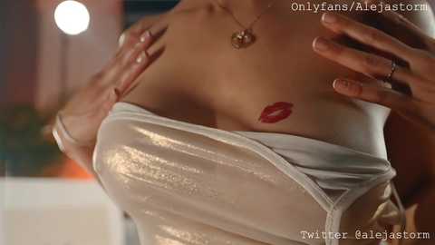 Media: Video of a woman's torso, with a see-through white dress revealing a red lipstick tattoo on her chest. The background is blurred, featuring warm lighting and a hint of greenery.