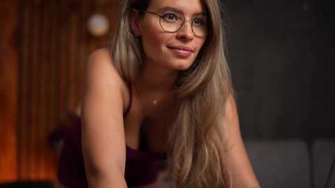 Media: Video of a Caucasian woman with long, straight blonde hair, wearing glasses and a purple top, leaning forward, smiling softly. Background features a blurred wooden wall and dim lighting.