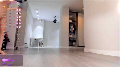 Media: Video of a modern, minimalist bedroom with light gray walls, wooden floor, and white furniture. A camera captures the scene from the corner, revealing a sleek, white desk and a closet with hanging clothes.