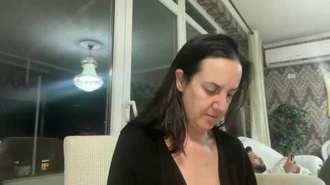 Media: Video of a woman with long black hair, wearing a black top, sitting indoors with a chandelier overhead, looking down.