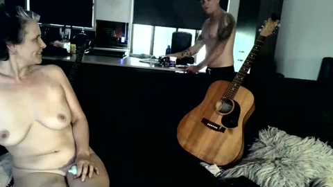 Media: Video of a naked man with a tattooed arm playing guitar, a nude woman with a tattoo on her side, and a wooden acoustic guitar on a black couch in a dimly lit room.