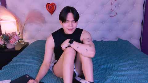 Media: Video of a smiling Asian woman with short black hair, wearing a black sleeveless top and shorts, sitting on a bed with a turquoise blanket, heart-shaped wall decor, and a lamp on a nightstand.