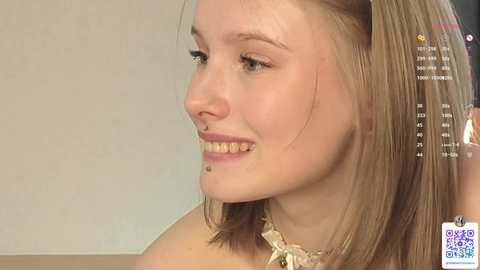 Media: Video of a young Caucasian woman with light skin, straight blonde hair, and a nose piercing. She has a warm smile and wears a lacy top. The background is a plain, off-white wall.