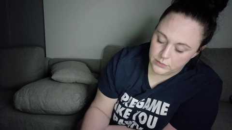 Media: A video of a pale-skinned woman with dark hair tied up, wearing a dark T-shirt with white text, sitting on a gray couch against a plain wall.