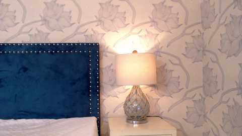 Media: Video of a bedroom corner with a dark blue velvet headboard, a white bedside table, and a glass lampshade with a floral design, against a white wallpaper with grey floral patterns.