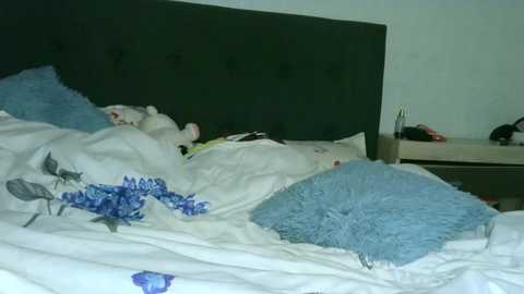 Media: Video of a messy bedroom with a dark green upholstered headboard, a plush white teddy bear, blue and white floral pillow, and a fuzzy light blue pillow on a white bedspread.