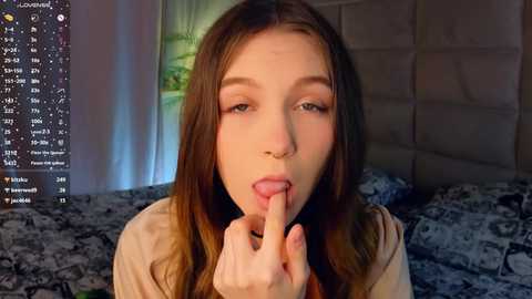 Media: Video of a young Caucasian woman with long brown hair, wearing a beige top, licking her finger suggestively, with a bed and digital screen in the background.