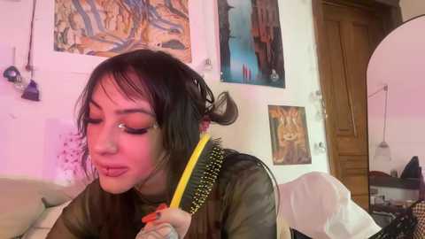 Media: Video of a young Asian woman with long black hair in a messy bun, using a yellow comb with black bristles to style her hair, surrounded by colorful art and wooden furniture in a cozy room.