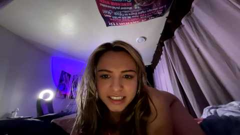 Media: Video of a young, light-skinned woman with long, straight brown hair, smiling, in a dimly lit bedroom with purple curtains and a neon light.