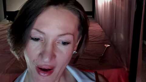 Media: A close-up video of a middle-aged woman with fair skin and brown hair, wearing a blue shirt, looking shocked and mouth agape. She is indoors on a bed with beige sheets, a metal object on the floor.