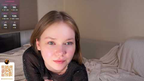 Media: Video of a young, fair-skinned girl with straight, light brown hair, wearing a sheer black top, lying on a bed with white sheets, in a softly lit bedroom.