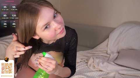Media: Video of a young, fair-skinned woman with long brown hair, wearing a black lace top, applying lotion to a man's exposed, tanned chest. They're in a dimly lit bedroom with beige walls.