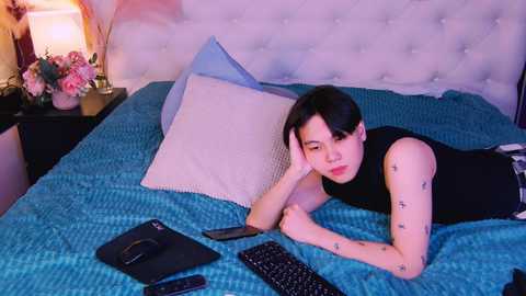 Media: Video of a young Asian woman with short black hair, lying on a blue-patterned bedspread in a dimly lit bedroom, wearing a black sleeveless top, with tattoos on her arms, using a smartphone and keyboard.