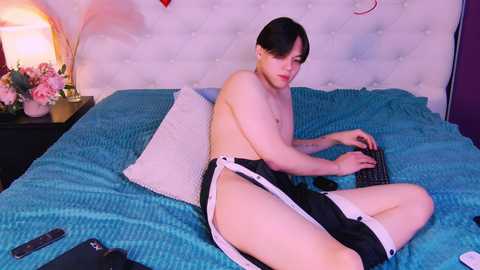 Media: Video of a young Asian man with short black hair, shirtless, wearing black shorts, playing a video game on a bed with blue sheets, surrounded by pink flowers, a lamp, and a remote control.