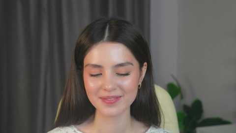 Media: Video of a young woman with long, straight black hair, fair skin, and light makeup, wearing a white lace top, smiling gently. Background features a gray curtain and a green plant.