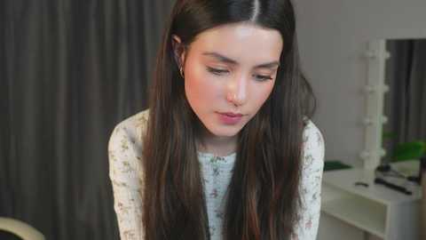 Media: Video of a young woman with long, dark hair, wearing a white floral top, and a nose ring, standing indoors against grey curtains and a white shelf.