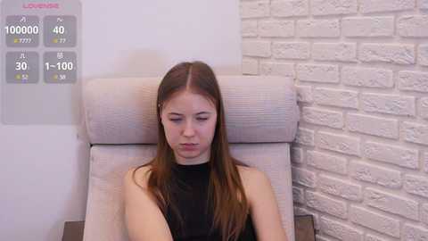 Video of a young, light-skinned woman with long brown hair, wearing a black sleeveless top, sitting in a beige armchair against a white brick wall. A digital display shows \"1000000\" in the background.