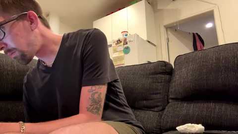 Media: A video of a bearded man in glasses, wearing a black T-shirt, leaning on a dark gray couch, with a tattoo on his right forearm. The background shows a white kitchen with a fridge and open door.