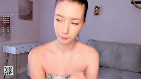 Media: A video of a fair-skinned, dark-haired woman with a slender physique, wearing a strapless snake-print dress, sitting on a grey couch in a minimalist, beige-walled room.