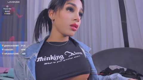 Media: Video of a Latina woman with light skin, black hair in a ponytail, wearing a black crop top and denim jacket, in a bedroom with white curtains and a bed.