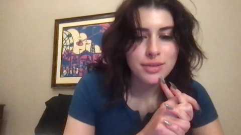 Media: Video of a young woman with dark, wavy hair, wearing a blue top, holding a finger to her lips. Background includes a framed, colorful painting and a beige wall.